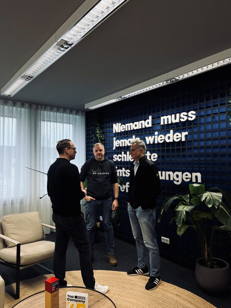 Mario Zillmann, Gero Presser und Christian Schneider im Gespräch in einem Büroraum. Im Hintergrund zieht man den Schriftzug "Niemand muss jemals wieder schlechre Entscheidungen treffen."